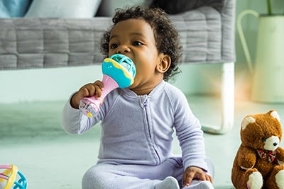 Black baby wearing purple onesie sitting on the ground with toy in their mouth