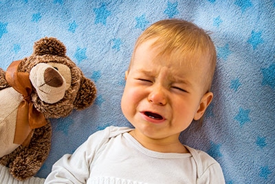 Crying Baby lying down against blue background with brown teddy bear