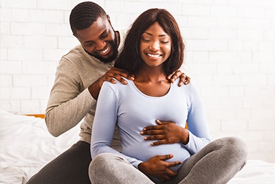 Black man massaging his pregnant wife shoulders, both sitting on bed at home