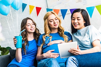 blonde pregnant young woman holding her belly is sitting between two brunette female friends looking at an ipad.