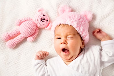 Yawning baby laying on white blanket wearing a pink hat next to a pink teddy bear.