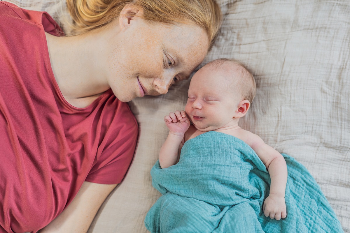A mother lying on a bed, looking lovingly at her sleeping newborn baby wrapped in a turquoise blanket.