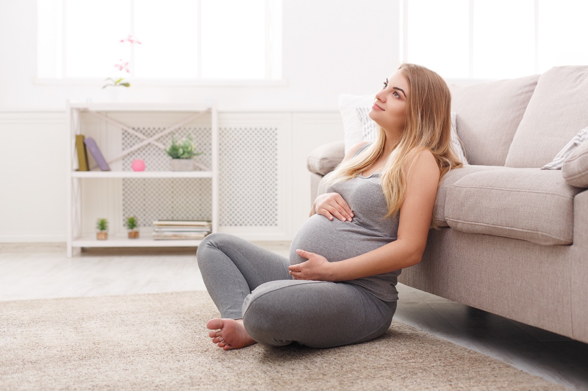 A pregnant woman sitting on the floor leaning against a sofa, smiling while resting her hands on her stomach